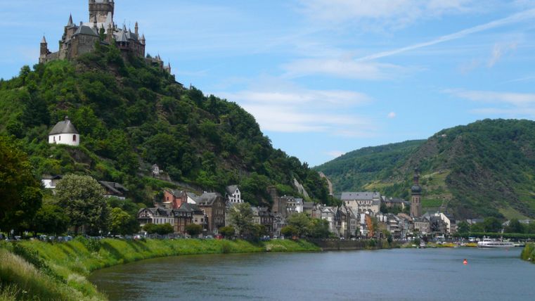 A picturesque landscape by the river with a castle on a hill. In the background, green mountains and blue sky can be seen.