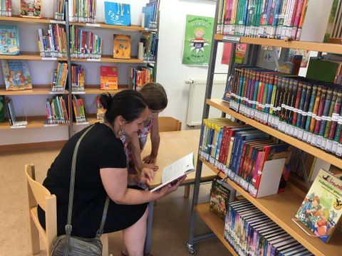 A woman and a child are sitting in a library, looking at a book together. In the background, there are shelves full of books.