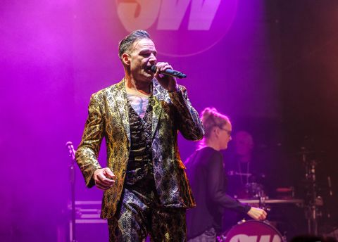 A musician in a striking golden suit is standing on stage and singing into the microphone. In the background, musicians and a colorful light show can be seen.