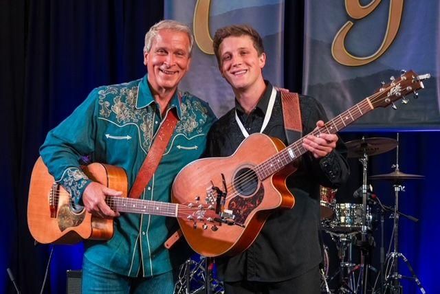 Zwei Musiker stehen lächelnd auf einer Bühne. Beide halten Akustikgitarren und tragen lässige Kleidung.