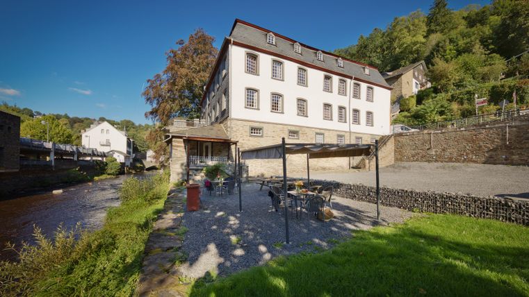 Ein Gebäude am Ufer eines Flusses, umgeben von Bäumen. Der Bereich vor dem Gebäude hat eine Terrasse mit Gartenmöbeln.