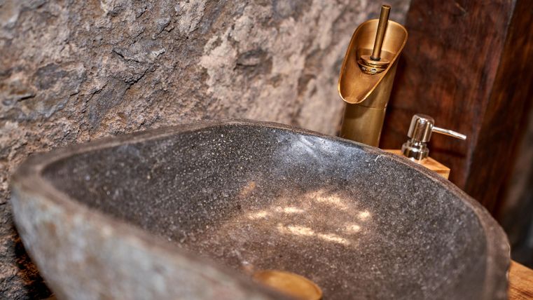 Ein elegenes Waschbecken aus dunklem Stein mit goldenen Armaturen. Die rustikale Umgebung aus Steinen und Holz ergänzt das moderne Design.