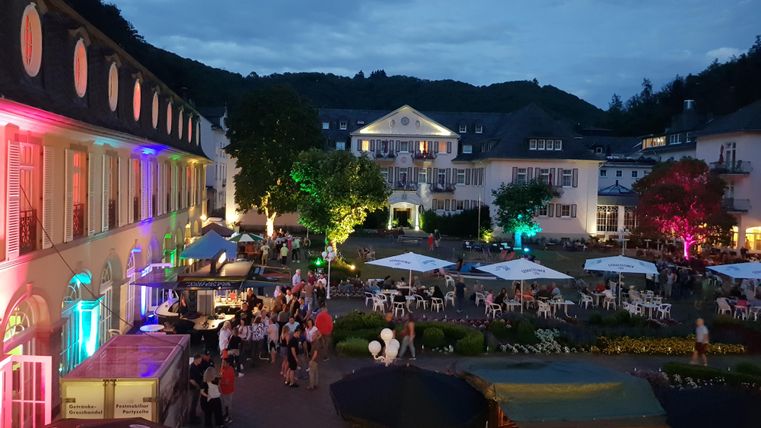 Een levendig plein 's nachts met kleurrijke lichten en veel mensen. Omgeven door historische gebouwen en zitruimtes buiten.