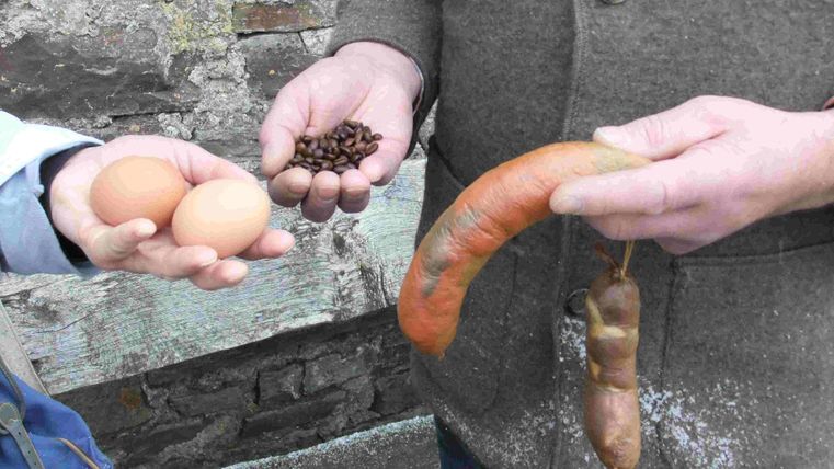 Two hands hold different foods: in one hand there are two eggs and in the other are coffee beans and a sausage. The scene shows a connection to rural nutrition.