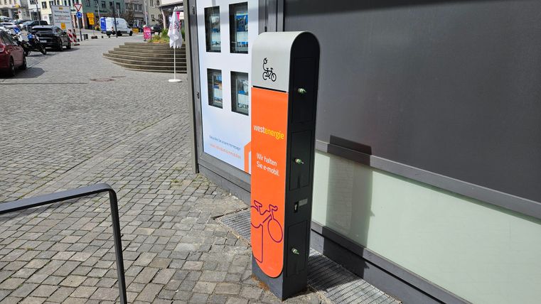 A bike station with an orange information sign is located on a paved path. In the background, several buildings and parked cars can be seen.