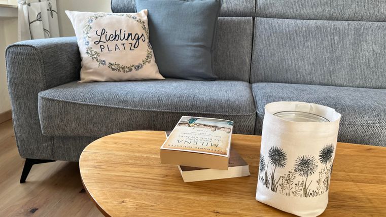 Ein gemütliches Wohnzimmer mit einem grauen Sofa und einem Kissen. Auf dem Tisch liegen Bücher und eine dekorative Lampe.