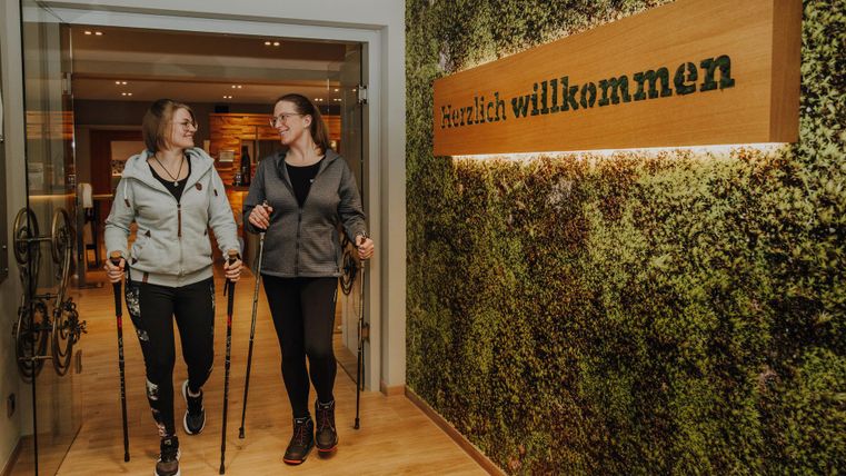 Twee vrouwen lopen met Nordic walking-stokken een vriendelijk ingerichte receptie binnen. Op de achtergrond hangt een welkomstbord aan een groene wand.