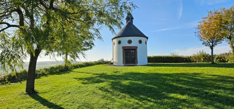 Auf einem Plateau steht in der Mitte eine runder, weißer Bau  mit einem Schieferdach und einem kleinem Kirchturm. Über der dunkelbraunen Flügeltür sind in regelmäßigem Abstand,  kleine runde Fenster eingelassen.Die Kapelle ist von einer grünen Wiese umgeben. Rechts und links stehen Laubbäume. Im Hintergrund umschließt eine Hecke das Gelände.
