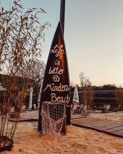 Ein Schild in Form eines Surfbretter zeigt die Botschaft: „Life is better @ Minden Beach“. Der Hintergrund ist eine entspannte Strandatmosphäre mit Sand und Pflanzen.