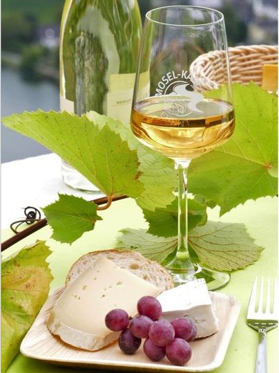 A glass of wine stands next to a cheese platter with grapes and fresh grape leaves. In the background, gentle hills and a water landscape can be seen.