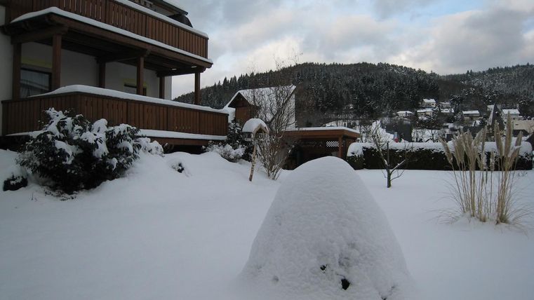 A snowy landscape with a house and a large snow mound in the foreground. In the background, snow-covered hills and trees can be seen.