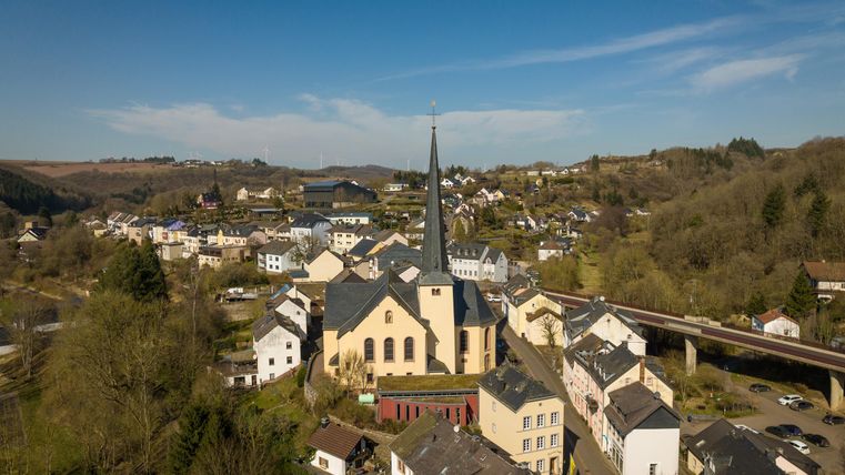 Eine malerische Dorfansicht mit einem Kirchgebäude im Mittelpunkt. Umgeben von Hügeln und Wohnhäusern, strahlt die Landschaft Ruhe aus.