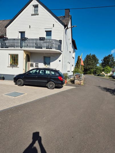 Ein modernes Wohnhaus mit einem Balkon und einem schwarzen Auto vor der Tür. Die Straße ist gut asphaltiert und lichtdurchflutet.