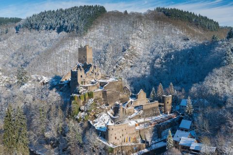 Eine alte Burgruine in einer schneebedeckten Landschaft. Im Hintergrund sind bewaldete Hügel zu sehen.
