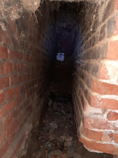 Un tunnel étroit, de couleur sombre, fait de briques avec des pierres et des débris au sol. Au bout du tunnel, une faible lumière est visible.