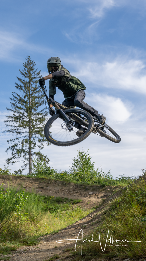Un cycliste de montagne saute avec son vélo par-dessus une colline. À l'arrière-plan, on peut voir des arbres et un ciel bleu.