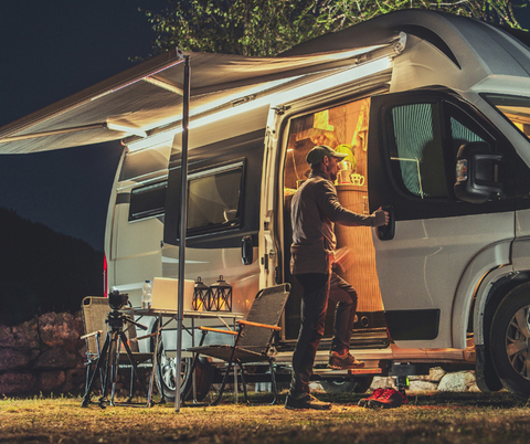 Une personne monte dans son camping-car. Dehors, il fait noir, la lumière à l'intérieur est allumée. Un pavillon est tendu sur le côté du camping-car.