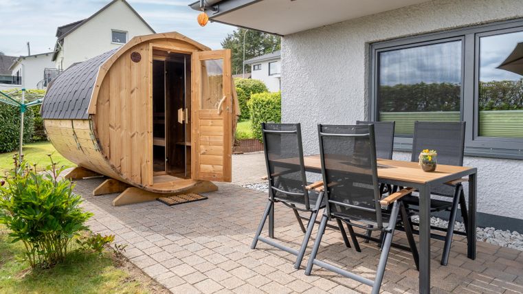 Ein schöner Gartenbereich mit einem Holzfass und einem Tisch mit Stühlen. Der Bereich ist umgeben von grünen Pflanzen und einem hellen Himmel.