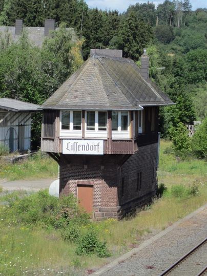Blick von oben auf ein altes etwa 3 Meter breites Gebäude.Das schmale  Erdgeschoss besteht aus roten Ziegelstein.Darauf befindet sich eine Holzkonstruktion aus braunen Brettern , die etwas breiter ist als das Erdgeschoss. Im unteren Bereich sieht man eine braune Tür. Der obere Bereich hat zur Front drei weiße Fenster. Unter den Fenstern ist ein weißes Schild mit der Aufschrift Lissendorf angebracht. Das Dach des Stellwerks ist mit schwarzem Schiefer eingedeckt.