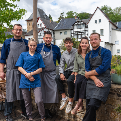 Six chefs are smiling in front of a background of historical buildings. It is a sunny day and the ambiance is inviting.