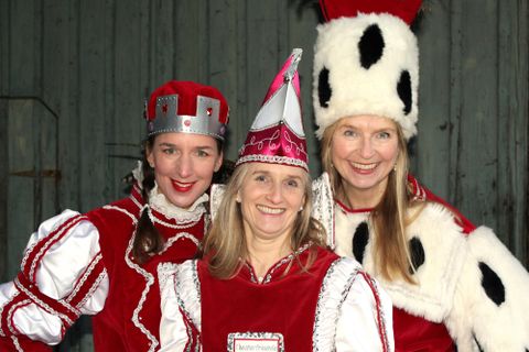 Three women in festive costumes smile at the camera. The outfits are red and white, with striking hats and a prince's crown.