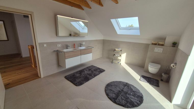 Une salle de bains moderne avec une décoration claire et des détails en bois. Il y a un double lavabo, des toilettes et une grande baie vitrée pour beaucoup de lumière.