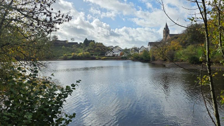 Ein ruhiger See umgeben von Bäumen und buntem Herbstlaub. Im Hintergrund sind Häuser und eine Kirche sichtbar.