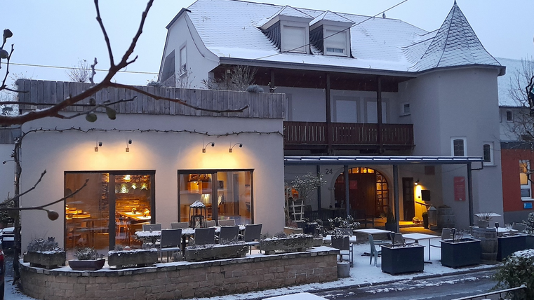 Ein einladendes Weinrestaurant mit hübscher Weinrebe im Vordergrund. Im Hintergrund sieht man das Restaurant und eine schöne Landschaft mit Weinbergen.
