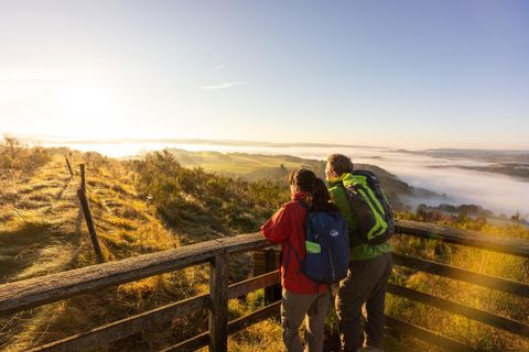 Een paar staat op een uitkijkplatform en kijkt naar een mistig landschap in het ochtendlicht. Het landschap toont zachte heuvels en een heldere lucht.