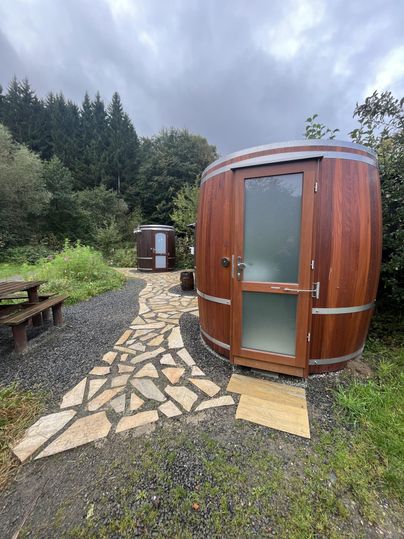 Deux tonneaux de sauna habillés de bois se trouvent dans un environnement naturel. Un chemin pavé mène aux tonneaux, entourés d'arbres verts et de buissons.