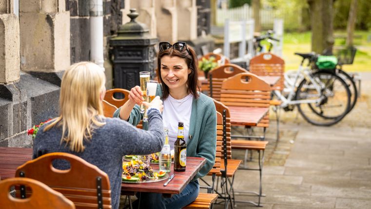 Twee vrouwen proosten met een drankje aan een tafel buiten, fietsen op de achtergrond.
