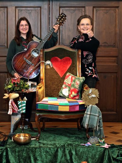 Zwei Personen stehen neben einem dekorierten Stuhl. Eine hat eine Gitarre in der Hand und beide lächeln fröhlich.