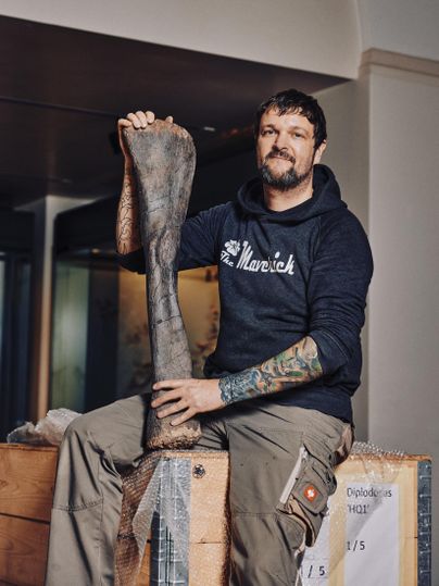 Ein Mann sitzt auf einer Holzkiste und hält ein großes Fossil in der Hand. Er trägt einen schwarzen Pullover und hat tätowierte Arme.