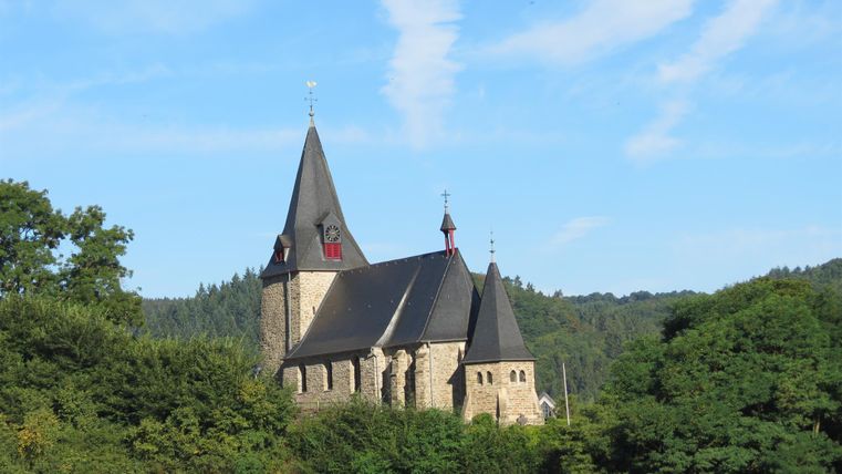 Eine alte Kirche mit einem markanten Turm steht umgeben von Bäumen. Der Himmel ist klar und blau mit einigen Wolken.