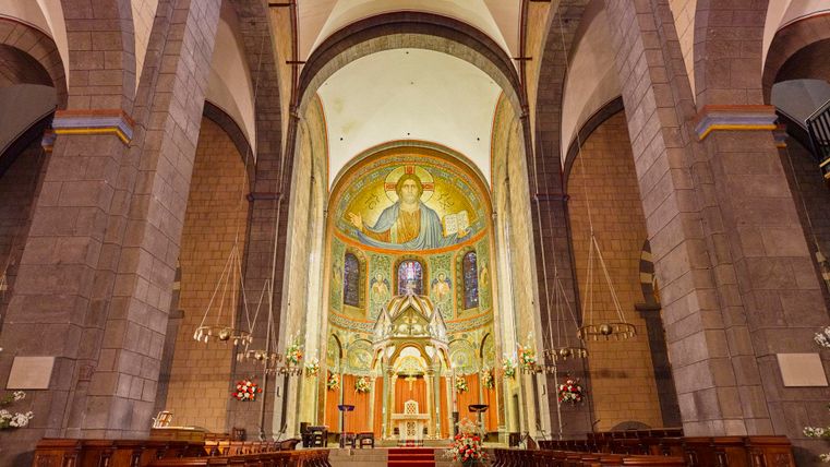 Een indrukwekkende kerk met hoge plafonds en kleurrijke muurschilderingen. Het rode tapijt leidt naar het altaar op de voorgrond.