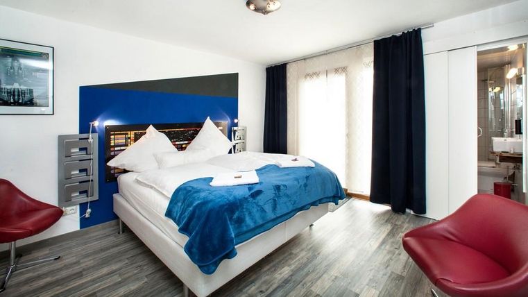 A modern bedroom with a large bed and blue accents. Red chairs complement the stylish decor, and the room has large windows for plenty of light.