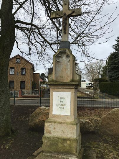 Une croix historique se dresse sur un socle en pierre. À l'arrière-plan, on aperçoit quelques bâtiments et des arbres.