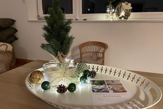 Ein dekorativer Tisch mit einem kleinen Weihnachtsbaum, einer Lichterkette und stilvollen Ornamenten. Auf einem Tablett liegen auch eine Zeitschrift und einige Tannenzapfen.