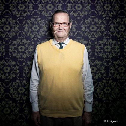 Een oudere man met een bril draagt een gele trui over een wit overhemd en een groene stropdas. De achtergrond is ingericht met een bloemmotief.