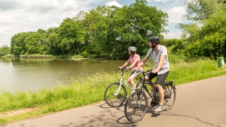 Zwei Personen fahren mit Fahrrädern auf einem Weg entlang eines Sees, umgeben von grünen Bäumen.