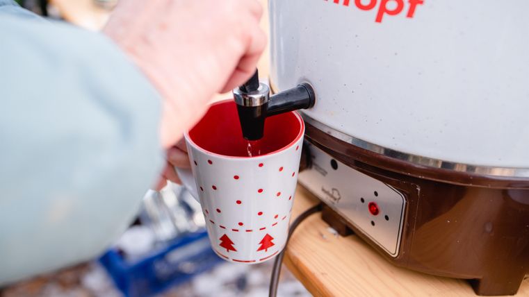 Een hand tapt een warme drank uit een drankdispenser in een rood-wit kopje. Het kopje is versierd met kerstmotieven.