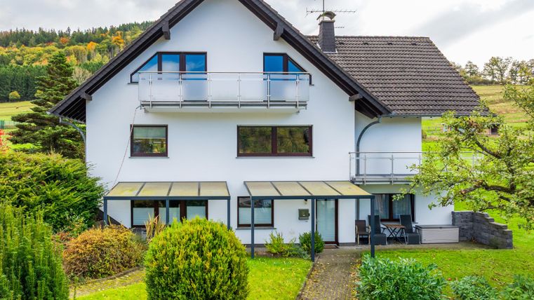 Une maison moderne avec un grand jardin. Elle a des balcons et est entourée d'arbres.