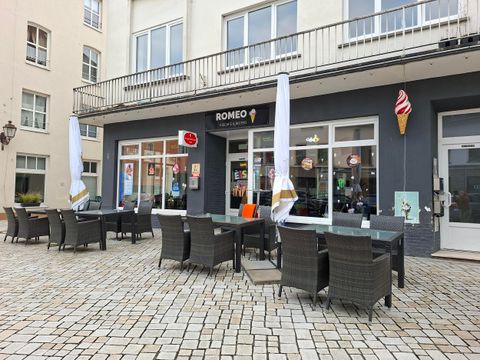 Ein Café mit Außensitzplätzen und Sonnenschirmen. Das Gebäude hat große Fenster und eine einladende Atmosphäre.