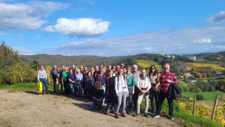 Eine Gruppe von Menschen steht auf einem Weg mit Blick auf eine malerische Landschaft. Der Himmel ist blau mit wenigen Wolken und die Umgebung ist grün und hügelig.