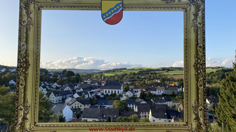 A picturesque view of a small town, framed by a golden picture frame. In the background, gentle hills and a clear sky can be seen.
