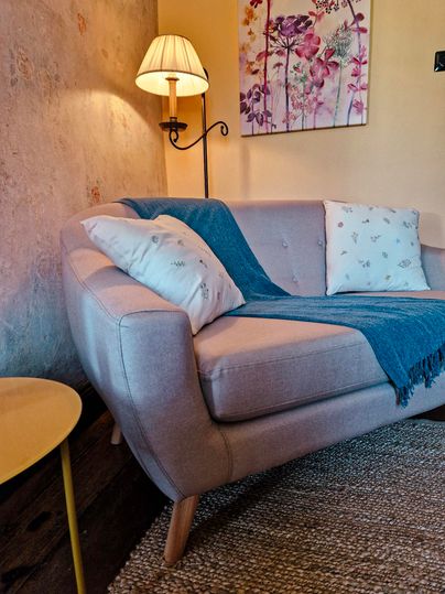 A cozy living room with a light sofa on which a blue blanket and cushions lie. Next to the sofa stands a small table, and a lamp provides warm light.