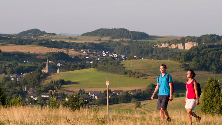 Two people walk through a hilly landscape with villages and fields in the background.