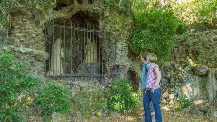 Twee mensen kijken naar een grot in de rots met beelden die beschermd worden door een traliewerk.