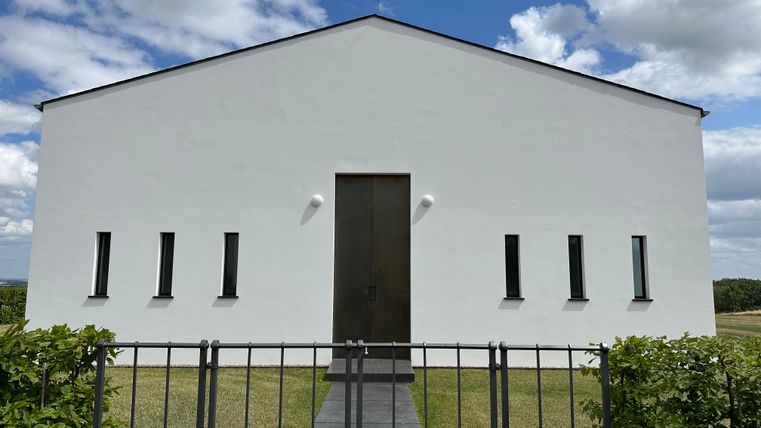 Bâtiment moderne blanc avec des fenêtres étroites et un grand portail en bois, entouré de pelouse et d'une clôture, sous un ciel bleu avec des nuages.