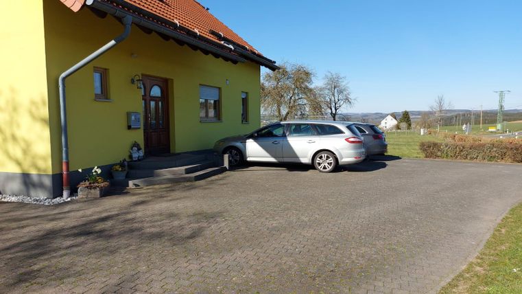 Een gele huis met een lichte lucht en een geplaveide oprit. Een zilveren auto staat voor de ingang.